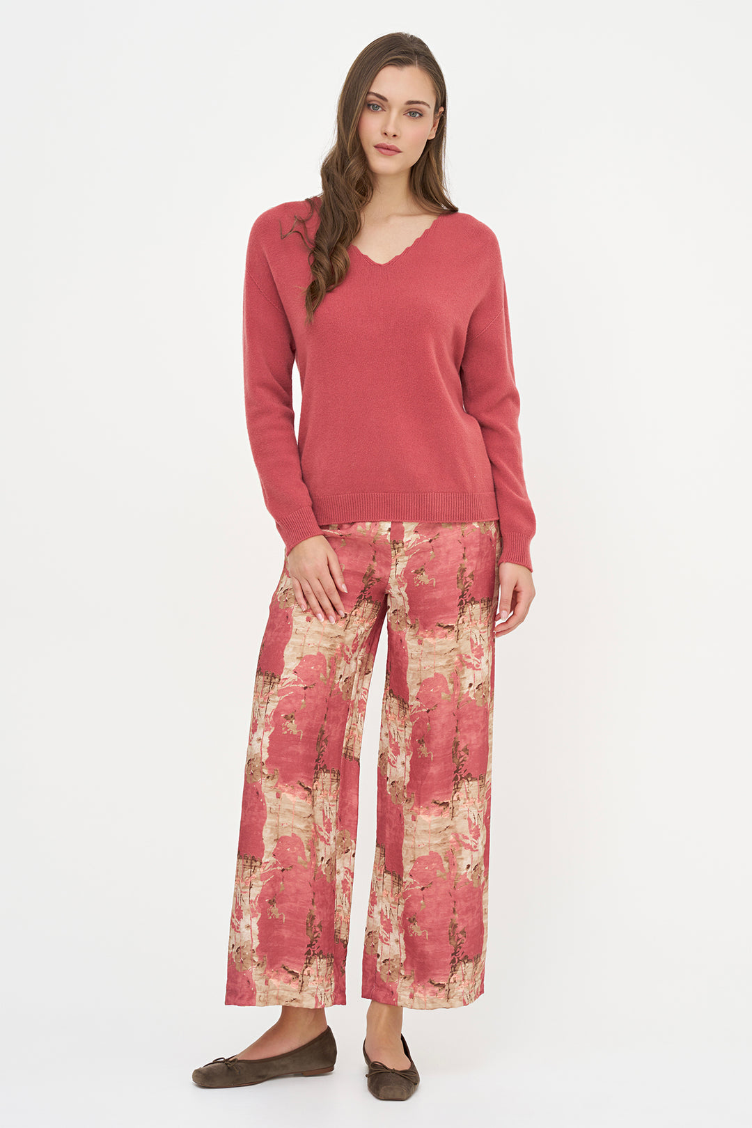 A woman modeling a two-piece outfit consisting of a solid dusty rose pink V-neck sweater and wide-leg trousers with a matching abstract floral print in pink, beige, and red tones, paired with dark brown flats against a plain white studio background.
