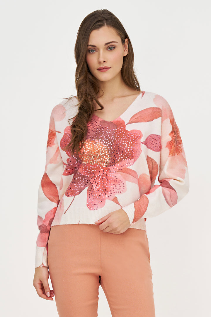 A woman modeling a long-sleeve V-neck top with a large pink and coral floral print over a white base, paired with peach pants, against a plain light gray studio background.