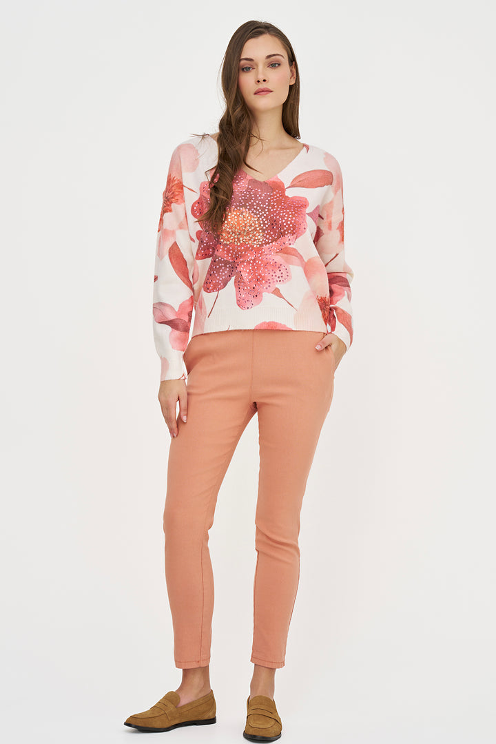A woman modeling a long-sleeve V-neck top with a pink and red floral print on a white background, paired with peach-colored slim-fit pants and brown loafers, against a plain white studio background.