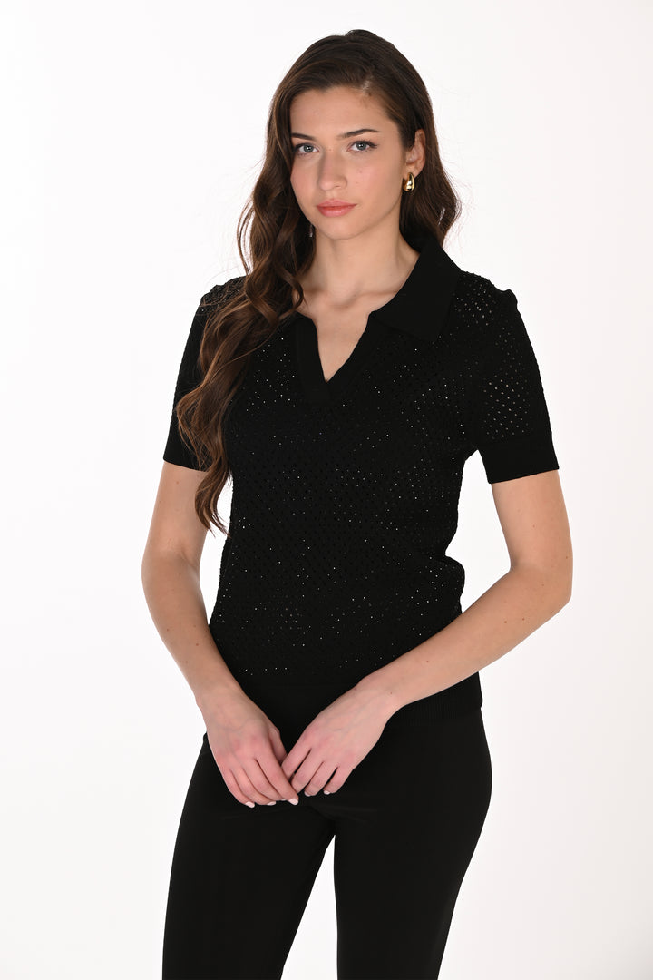 A woman modeling a black short-sleeve knit polo shirt with a V-neckline and subtle textured dot pattern, paired with black pants against a plain white studio background.