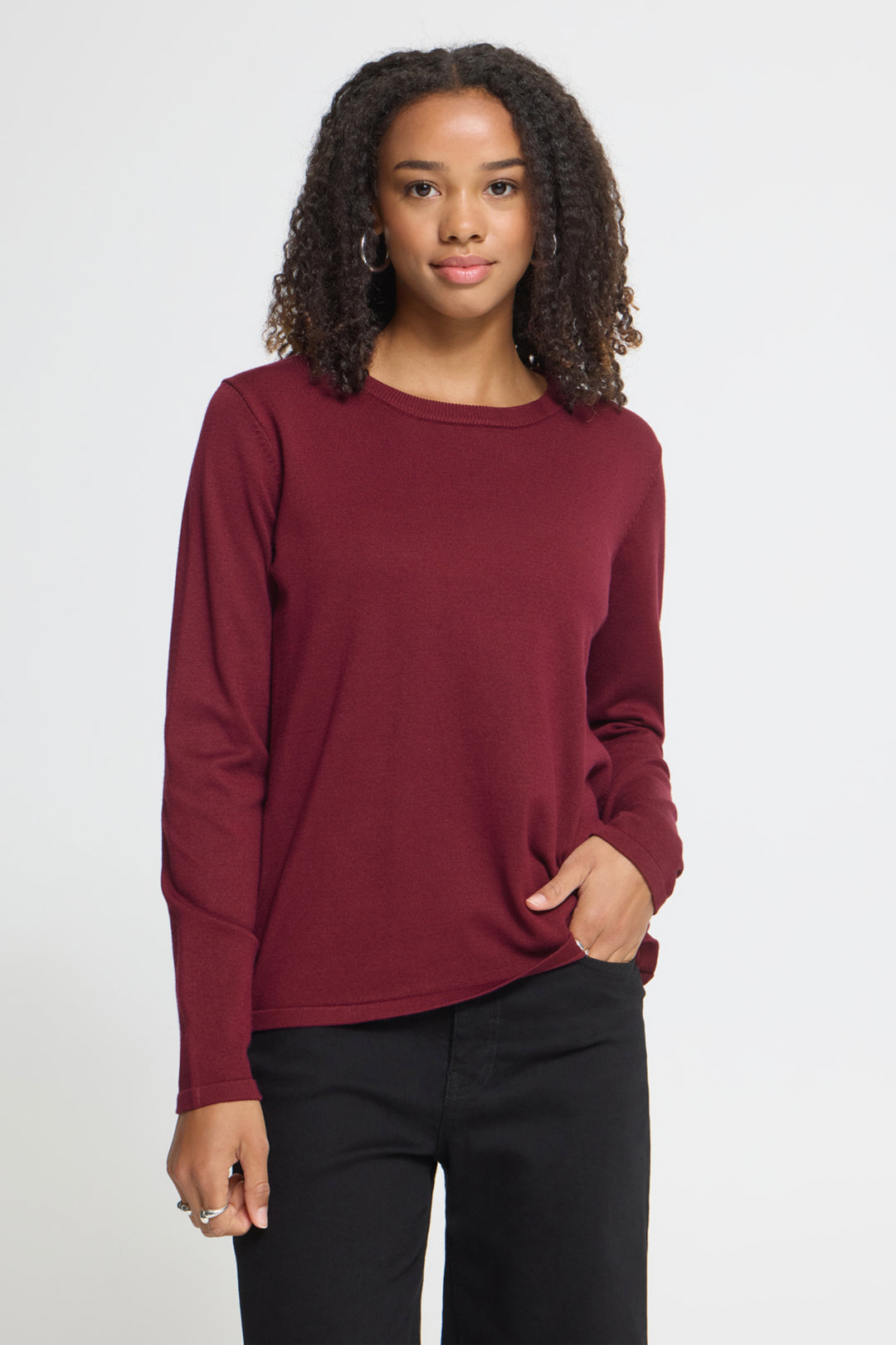 A woman with curly dark hair wearing a long-sleeve crewneck top in burgundy and black pants, standing against a light gray studio background.