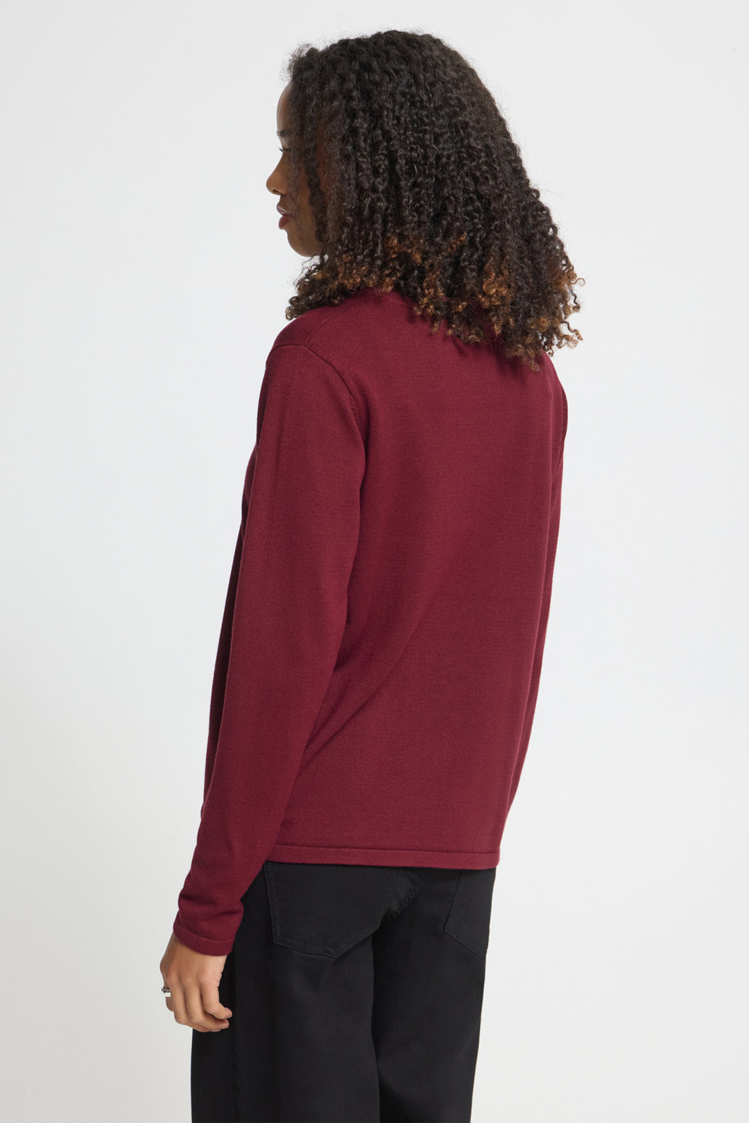 A woman with curly dark hair is shown from the back wearing a long-sleeve crewneck sweater in burgundy and black pants, photographed against a plain light gray studio background.