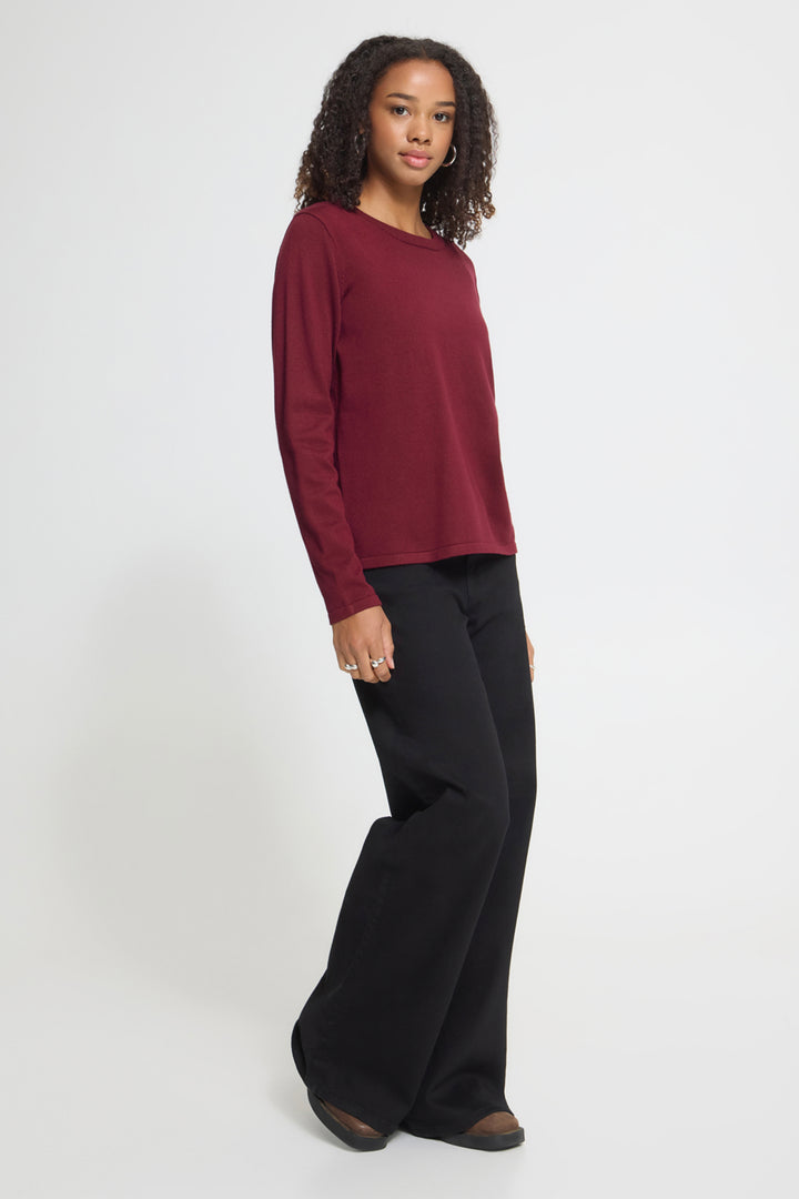 A woman modeling a long-sleeve crewneck top in deep burgundy paired with black wide-leg pants, standing against a light gray studio background.