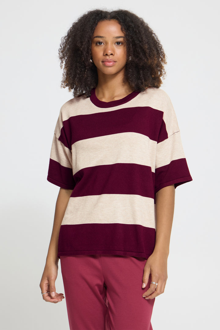 A woman with curly dark hair wearing an oversized short-sleeve striped t-shirt with horizontal bands of deep burgundy and cream, paired with dusty rose pants, against a light gray studio background.