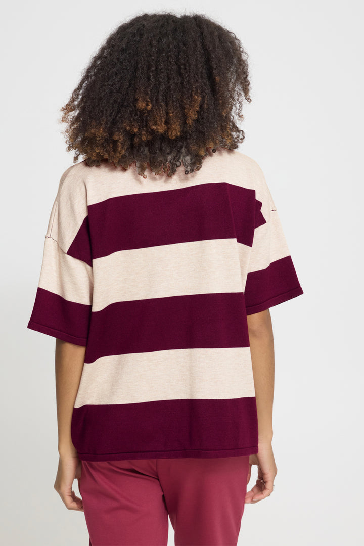 A person shown from the back wearing an oversized short-sleeve striped top with wide cream and deep burgundy horizontal bands, paired with burgundy pants, against a light gray studio background.