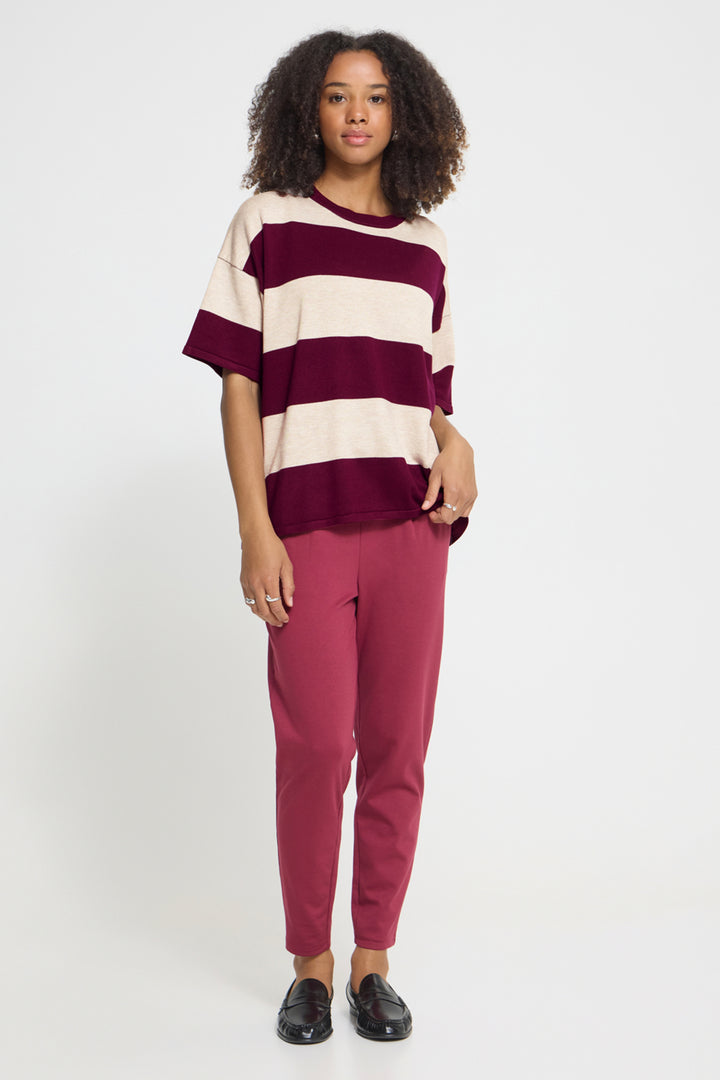 A woman modeling a short-sleeve knit top with wide burgundy and cream horizontal stripes, paired with dusty rose pants and black loafers, against a plain light gray studio background.