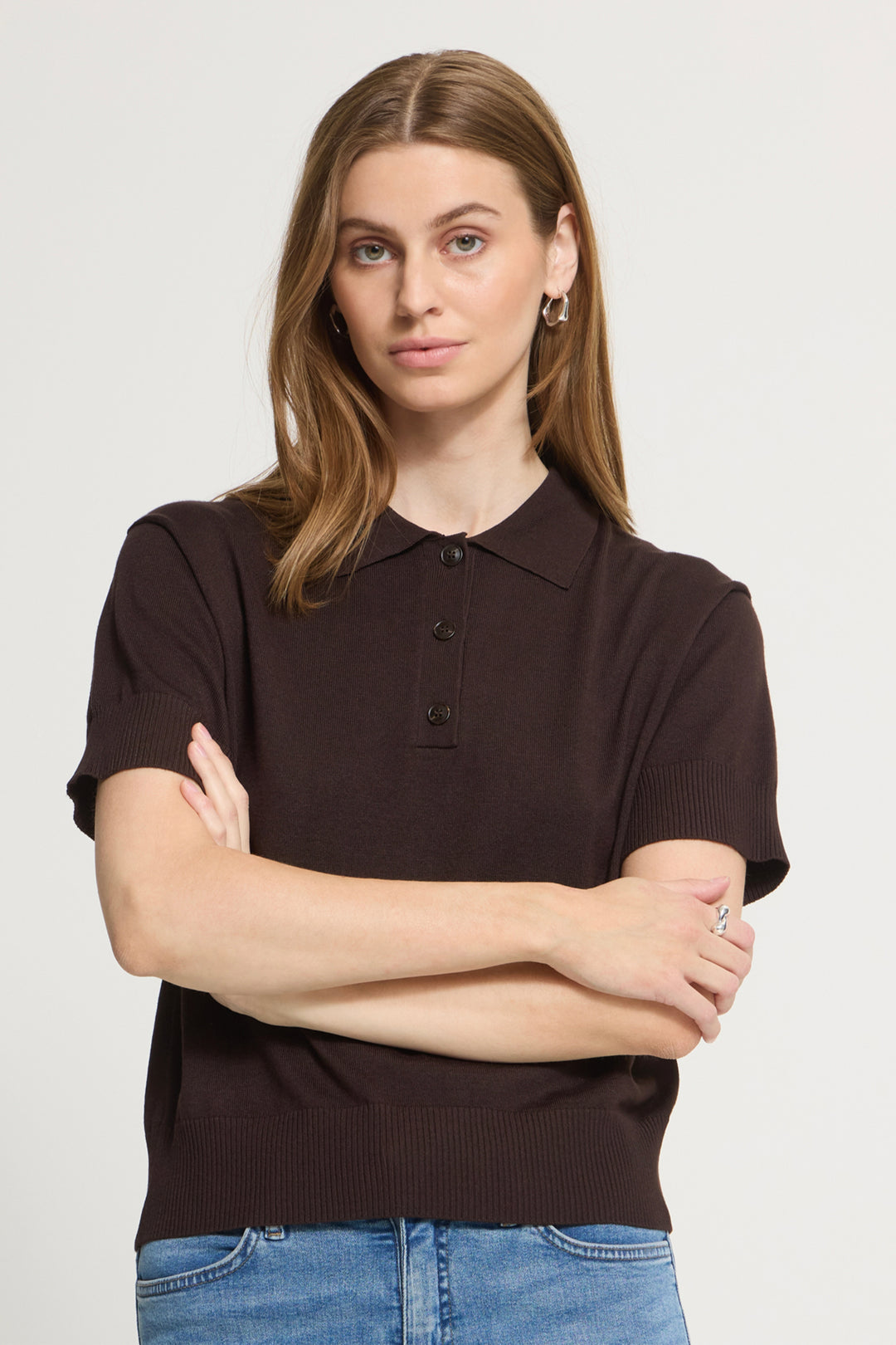 A woman wearing a dark brown short-sleeve knit polo shirt with blue jeans, standing against a light gray studio background with her arms crossed.