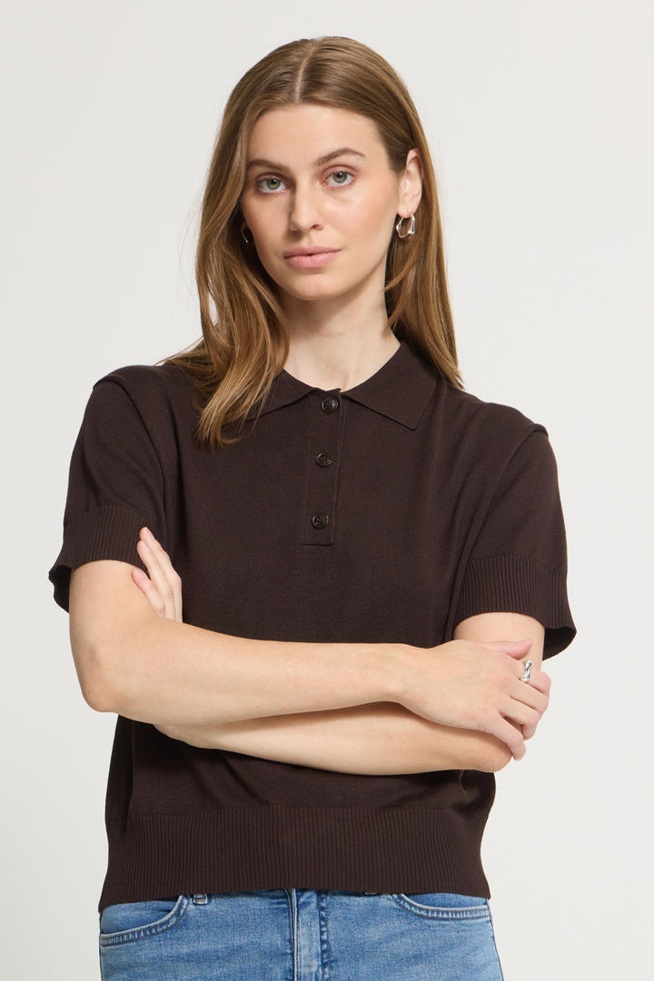 A woman wearing a dark brown short-sleeve knit polo shirt with blue jeans, standing against a light gray studio background with her arms crossed.