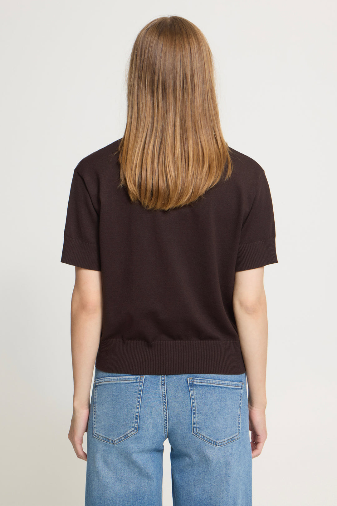 A woman viewed from the back wearing a dark brown short-sleeve knit top and light blue jeans against a plain light gray studio background.
