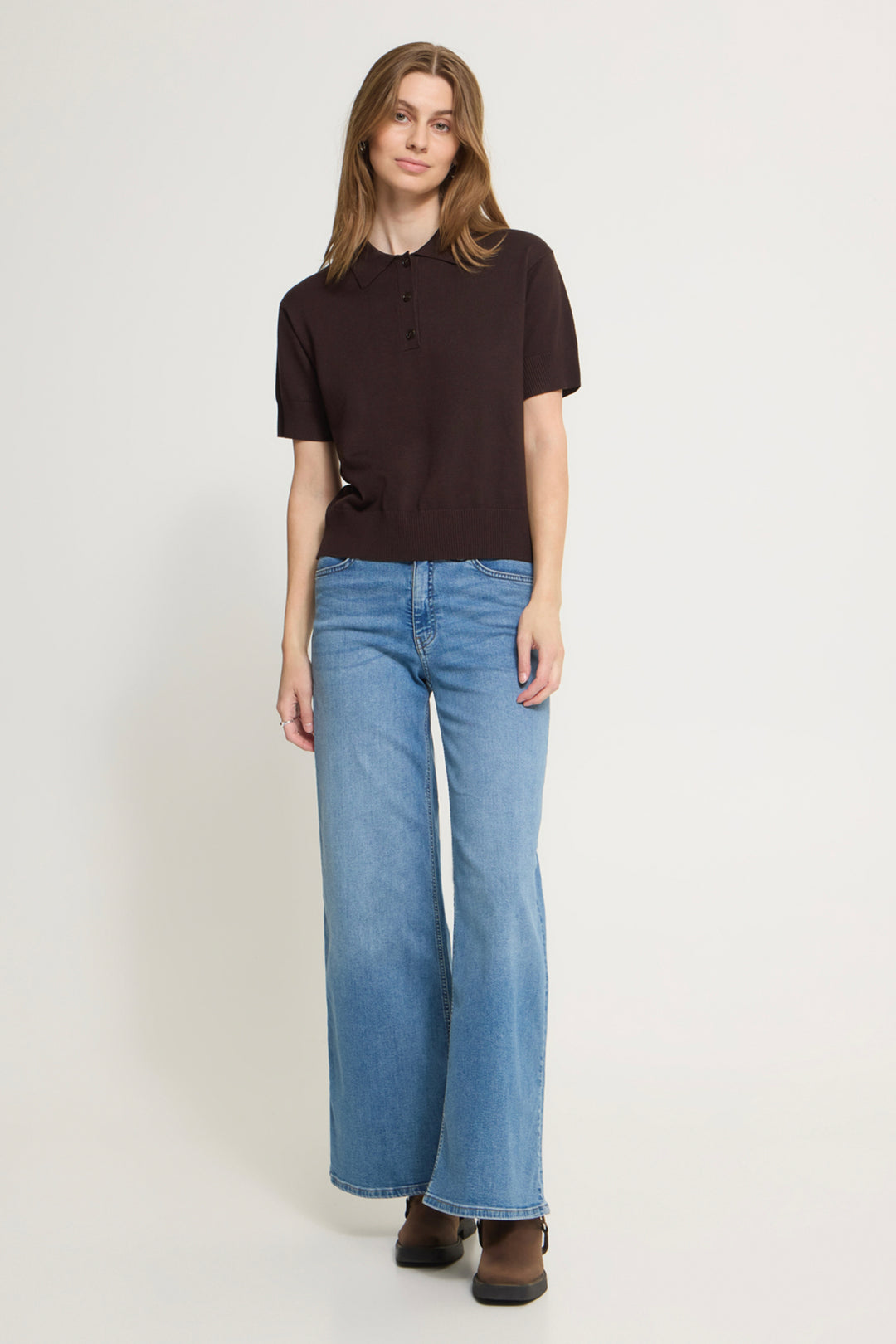 A woman modeling a dark brown short-sleeve henley top and light blue wide-leg jeans against a plain light gray studio background.