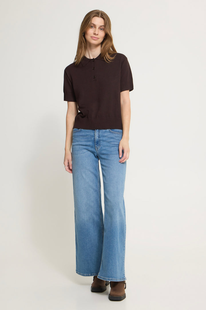 A woman modeling a dark brown short-sleeve henley top and light blue wide-leg jeans against a plain light gray studio background.