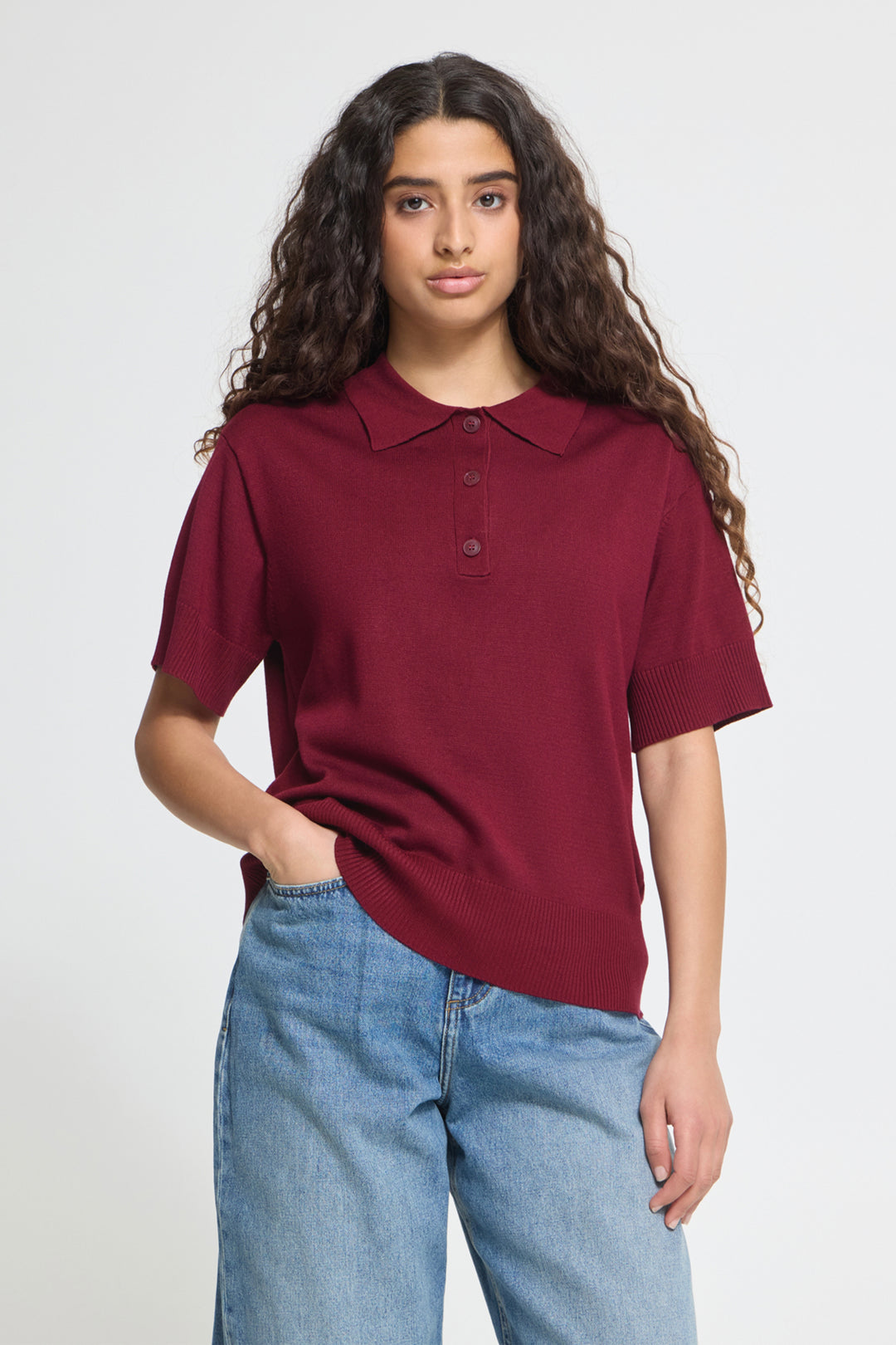 A woman with long, curly brown hair wearing a short-sleeve polo shirt in maroon and light wash blue jeans, standing against a light gray studio background.