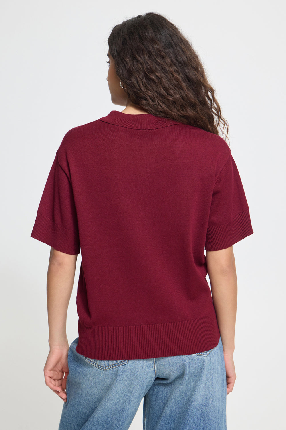 A woman viewed from the back wearing a short-sleeve knit top in deep burgundy and light blue jeans, against a plain light gray studio background.
