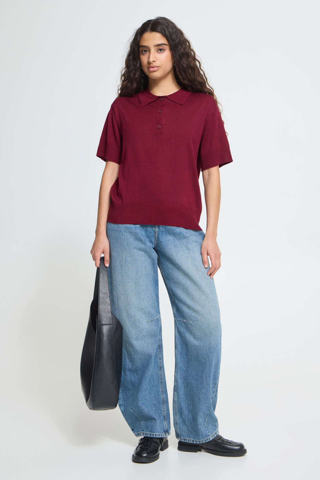 A woman modeling a short-sleeve polo shirt in burgundy, paired with wide-leg light blue jeans and black shoes, holding a black shoulder bag against a plain light gray studio background.