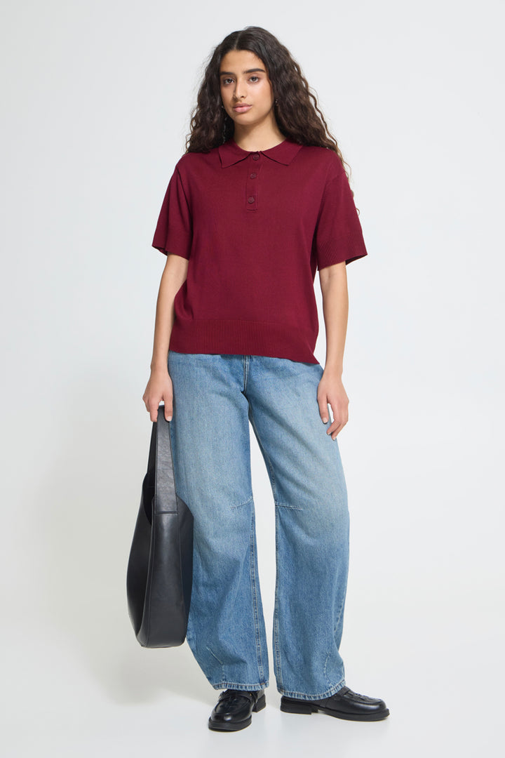 A woman modeling a short-sleeve polo shirt in burgundy, paired with wide-leg light blue jeans and black shoes, holding a black shoulder bag against a plain light gray studio background.