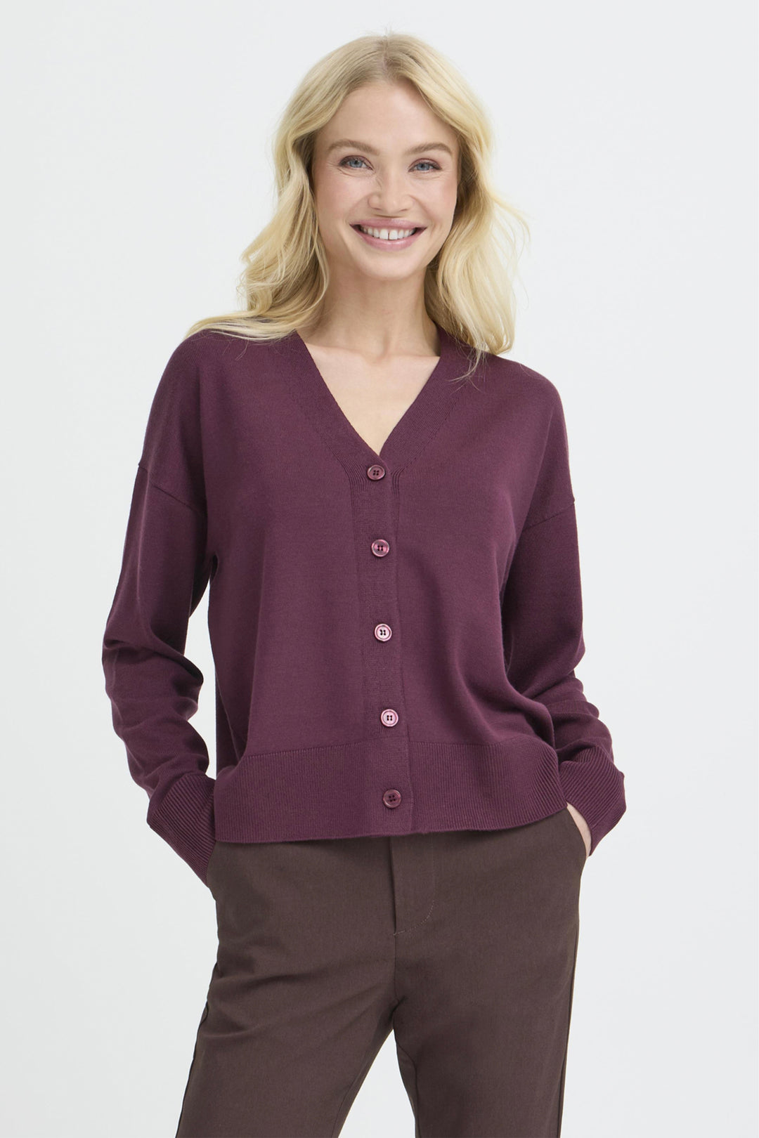 A smiling blonde woman wearing a long-sleeve V-neck cardigan in deep plum purple with matching buttons, paired with dark brown trousers, against a light gray studio background.