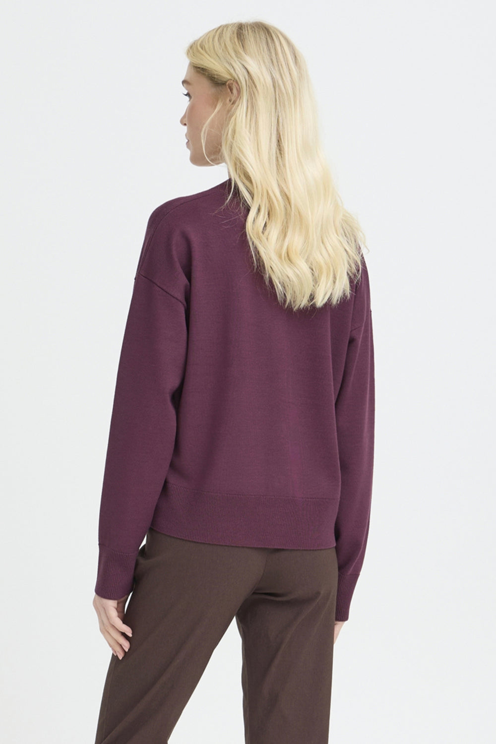 A woman with long blonde hair is shown from the back wearing a dark plum purple knit sweater and brown trousers against a light gray studio background.
