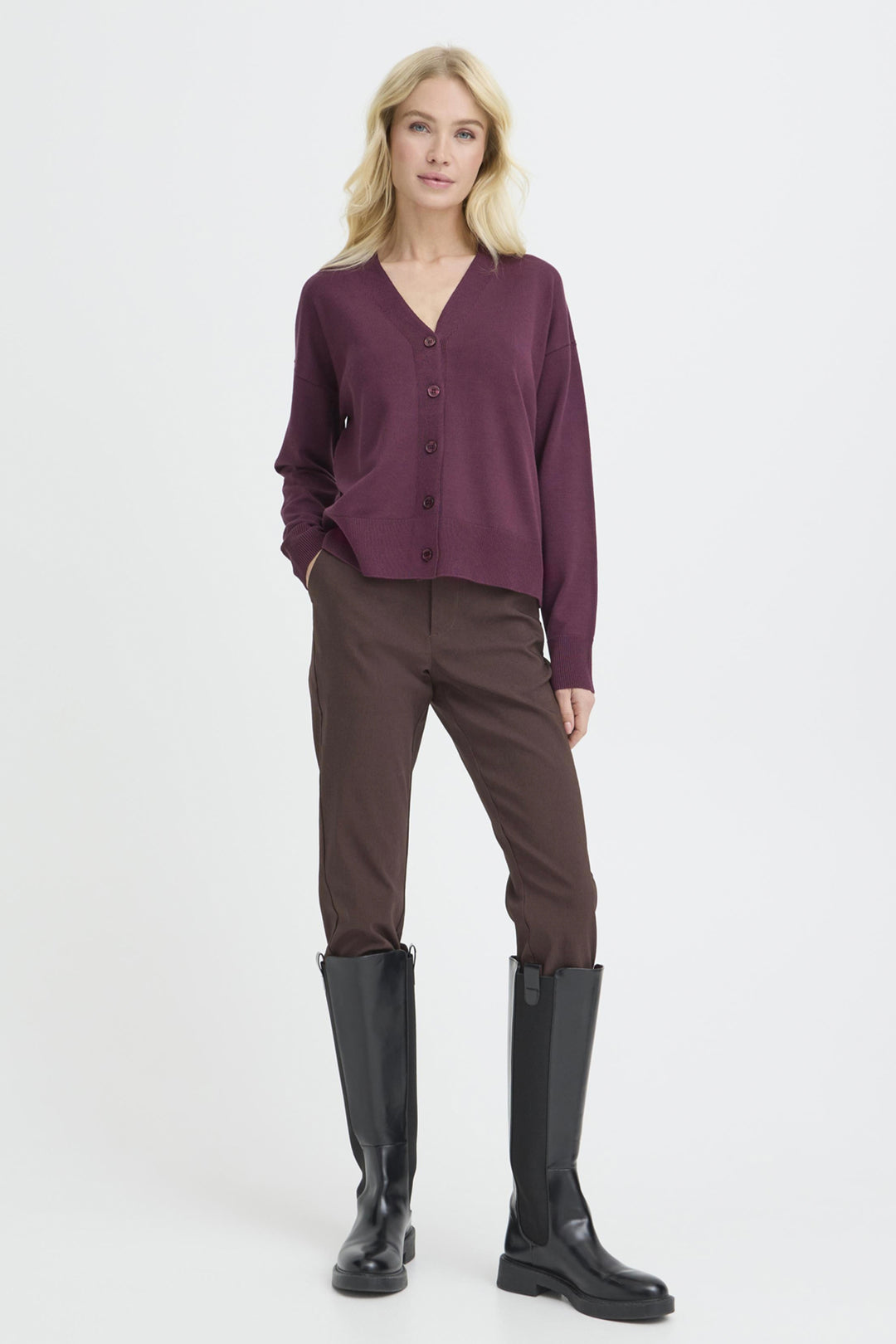 A woman modeling a long-sleeve button-front cardigan in deep plum purple, paired with dark brown trousers and black knee-high boots, against a plain light gray studio background.