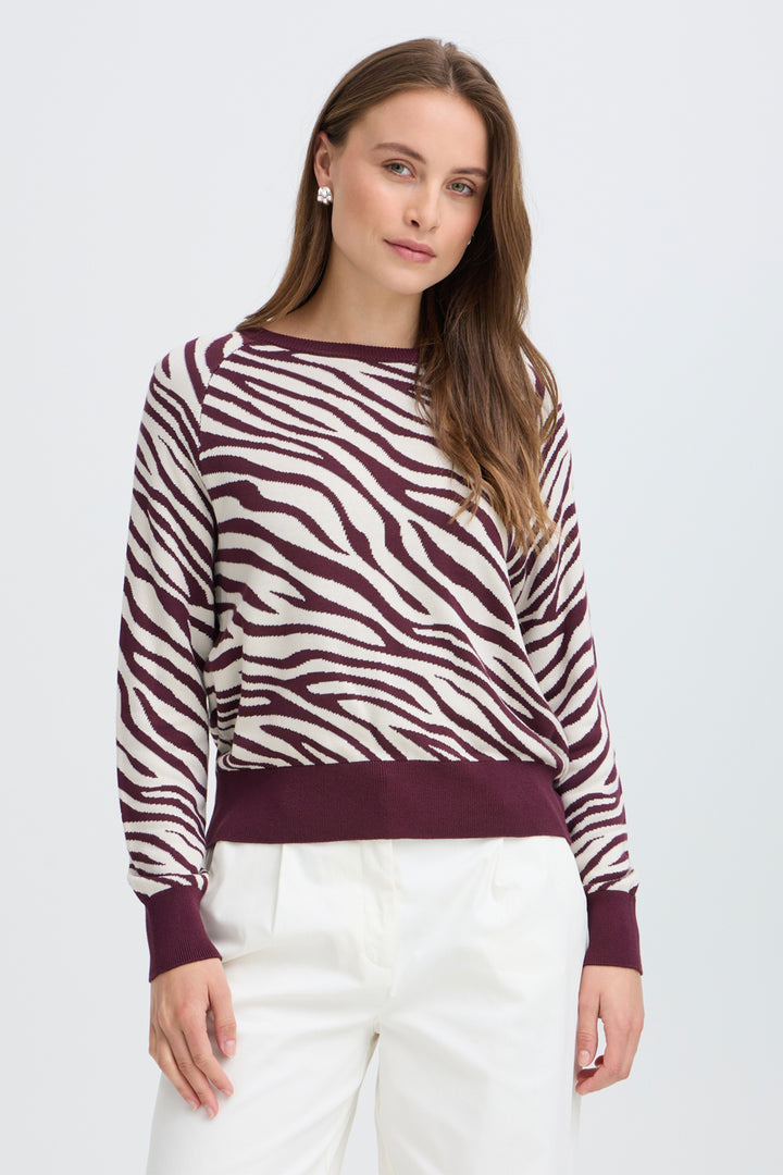 A woman modeling a long-sleeve knit sweater with a zebra-stripe pattern in burgundy and off-white, paired with white pants, against a light gray studio background.