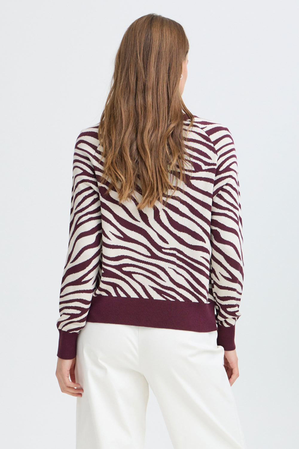 A woman viewed from the back wearing a long-sleeve knit top with a zebra stripe pattern in burgundy and off-white, paired with white pants, against a light gray studio background.