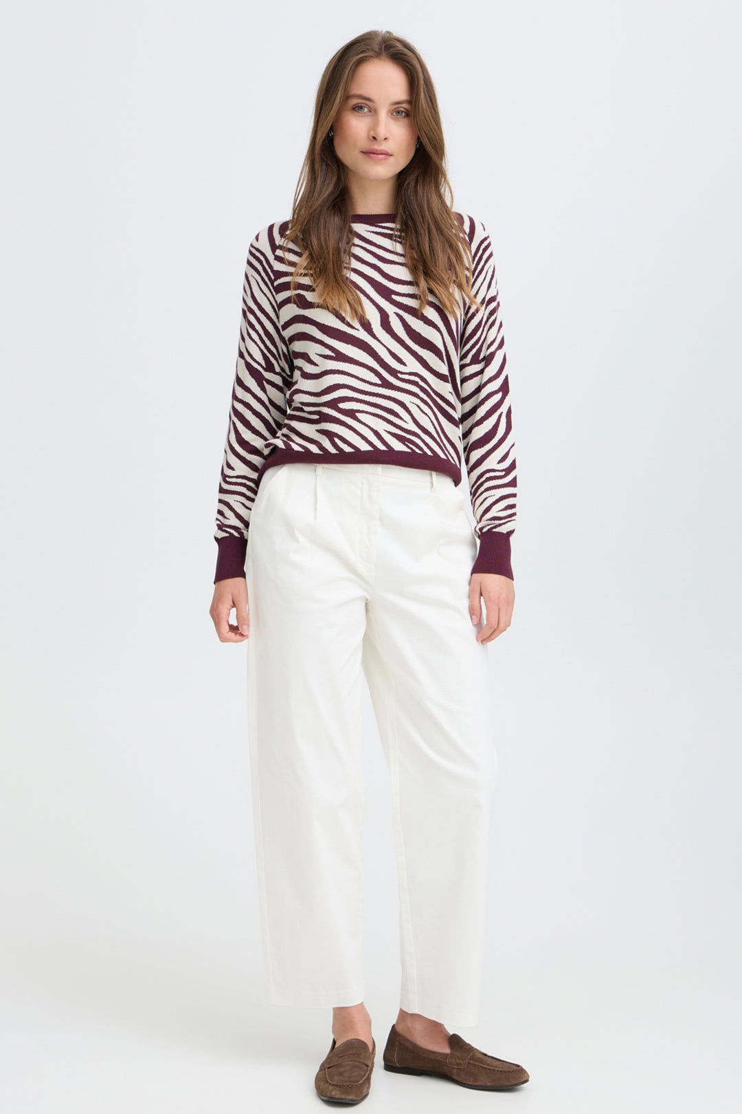A woman modeling a long-sleeve knit sweater with a brown and white zebra pattern, paired with white cropped trousers and brown suede loafers, against a plain white studio background.