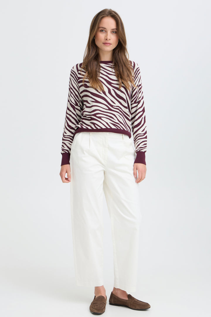 A woman modeling a long-sleeve knit sweater with a brown and white zebra pattern, paired with white cropped trousers and brown suede loafers, against a plain white studio background.