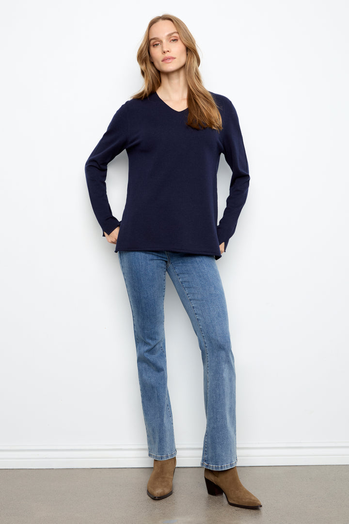 A woman modeling a navy blue long-sleeve V-neck knit top with light-wash blue jeans and brown ankle boots, standing against a plain white wall.