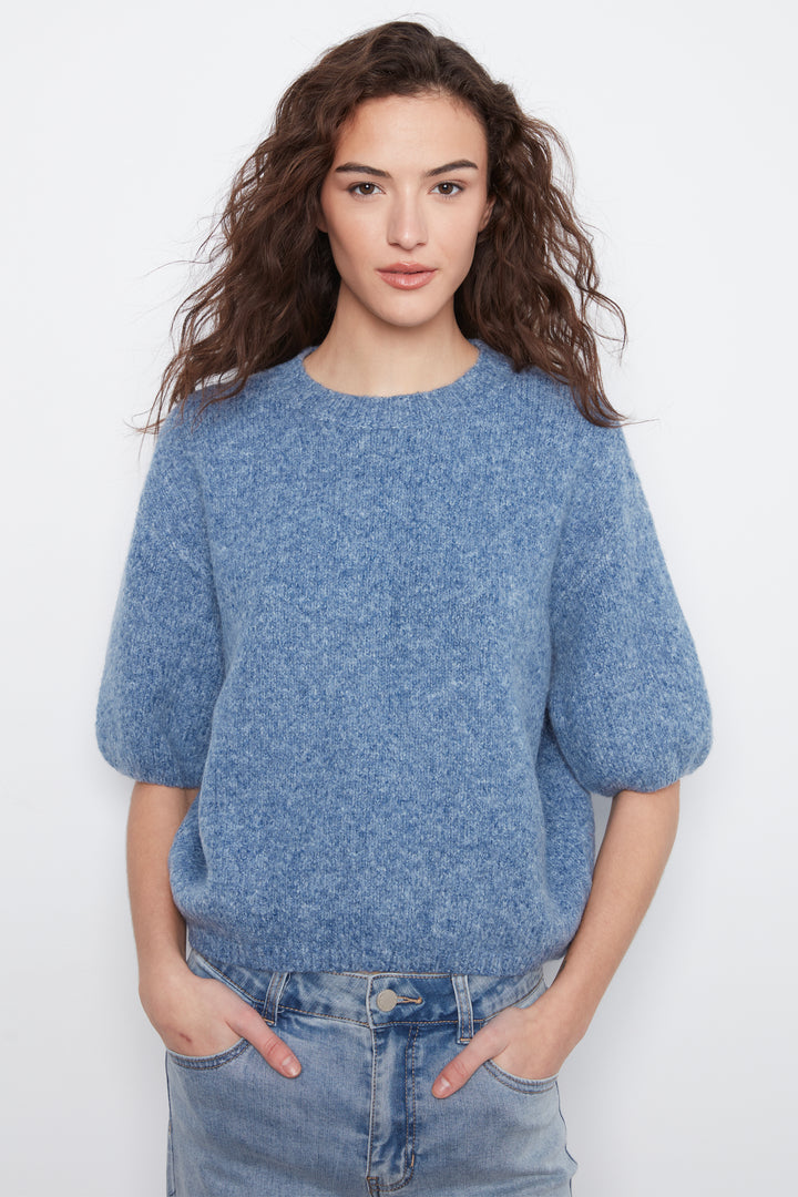 A woman with long, wavy brown hair wearing a heathered blue short-sleeve knit sweater and light-wash blue jeans, standing against a plain white studio background.