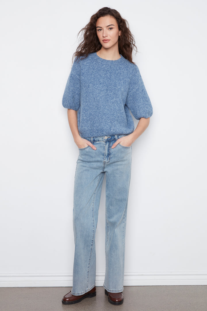A woman stands against a plain white wall wearing a heathered blue short-sleeve knit top, light-wash blue jeans, and dark brown shoes.