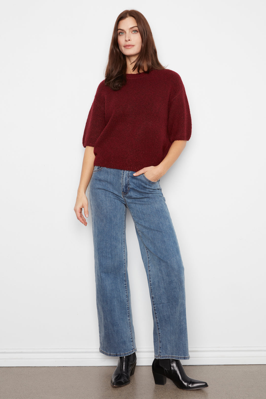 A woman modeling a dark red short-sleeve knit top and wide-leg blue jeans with black ankle boots, standing against a plain white wall.