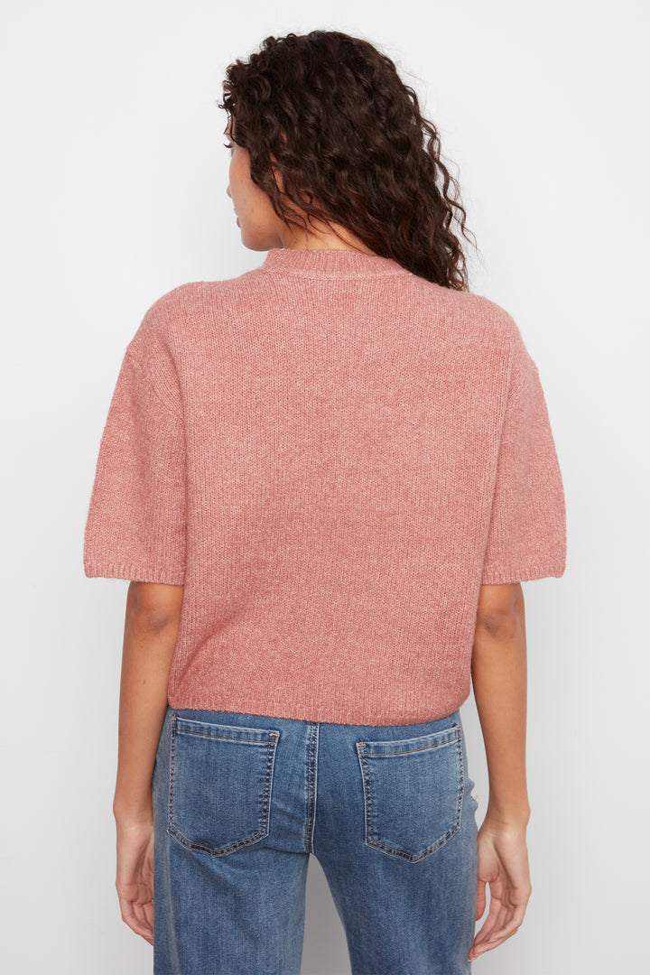 A woman viewed from the back wearing a dusty rose short-sleeve knit top and blue jeans against a plain light gray background.