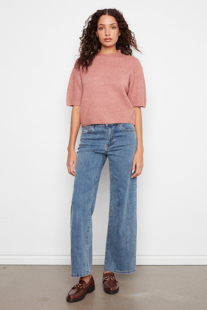 A model stands against a white wall wearing a dusty rose short-sleeve knit top, blue wide-leg jeans, and brown leather loafers.