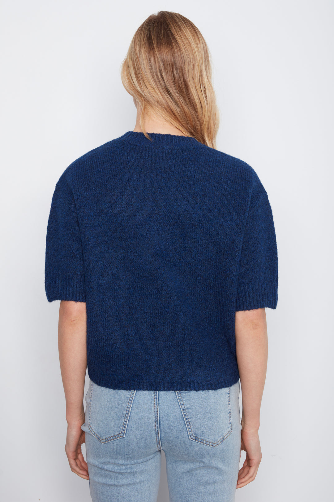 A woman viewed from the back wearing a navy blue short-sleeve knit sweater and light-wash blue jeans against a plain white background.