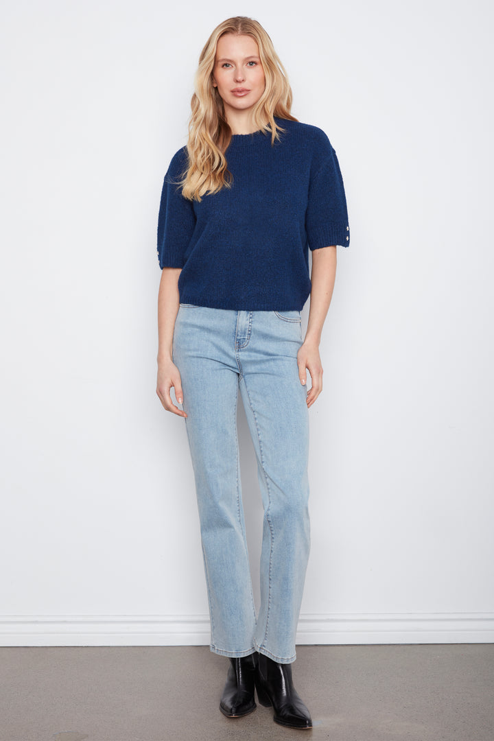 A woman stands against a plain white wall wearing a navy blue short-sleeve knit top, light-wash blue jeans, and black ankle boots.