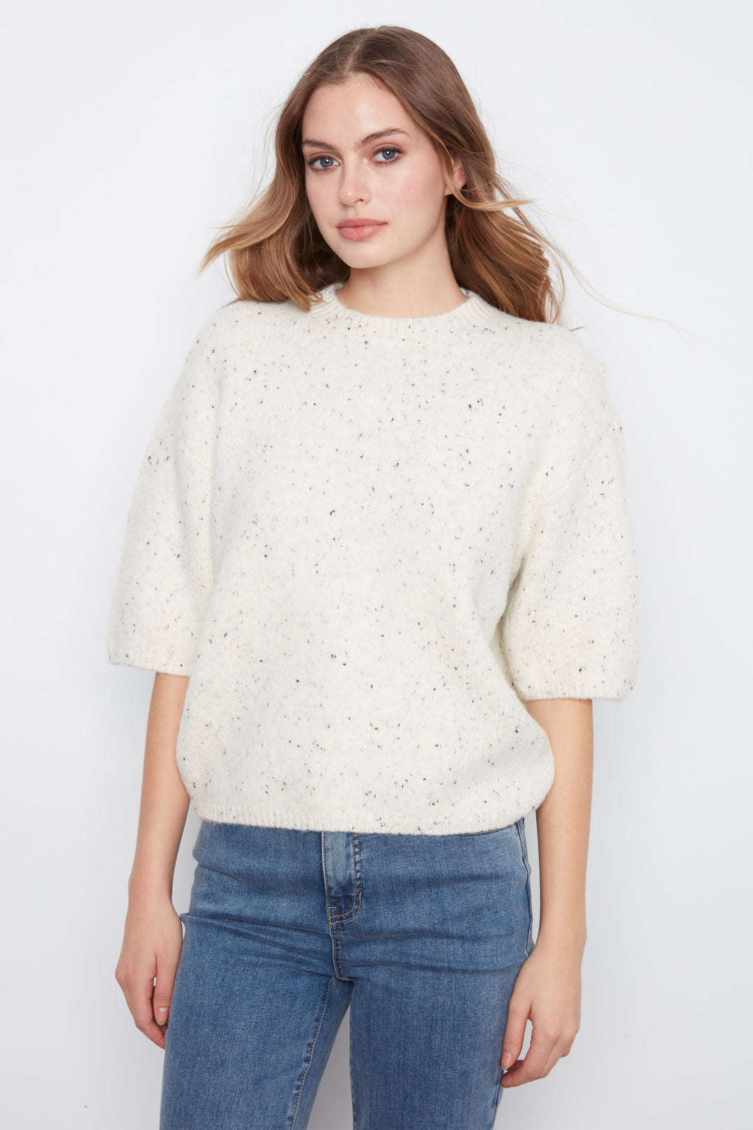 A woman wearing a cream-colored, textured knit sweater with small speckles and elbow-length sleeves, paired with blue jeans, against a plain white background.