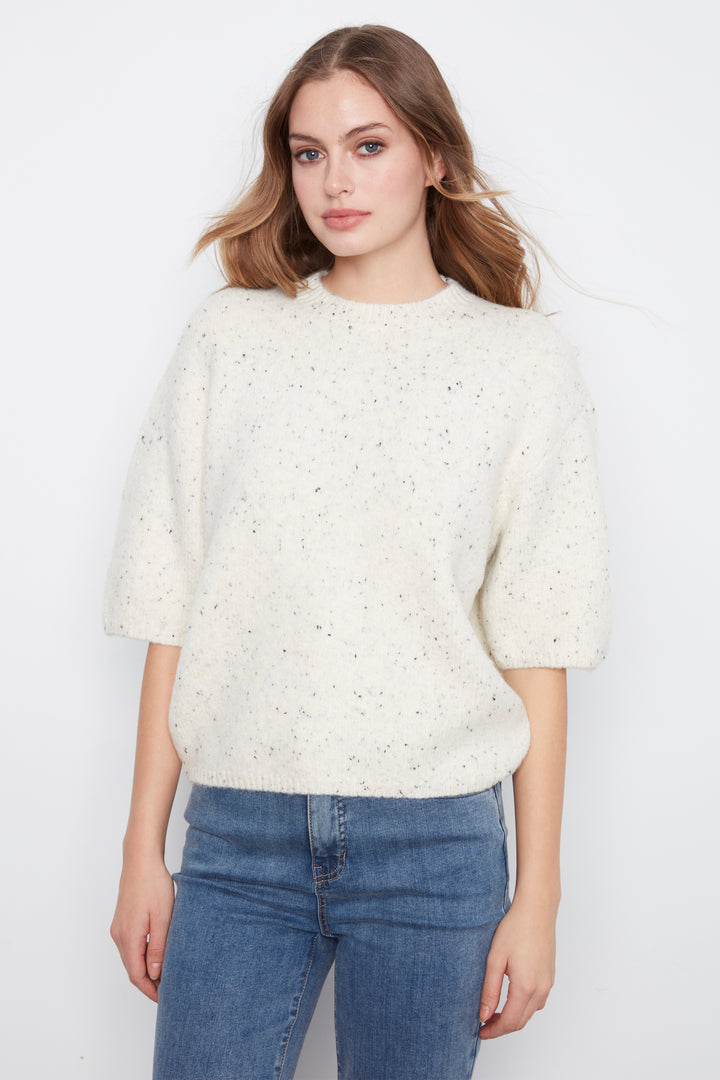 A woman wearing a cream-colored, textured knit sweater with small speckles and elbow-length sleeves, paired with blue jeans, against a plain white background.