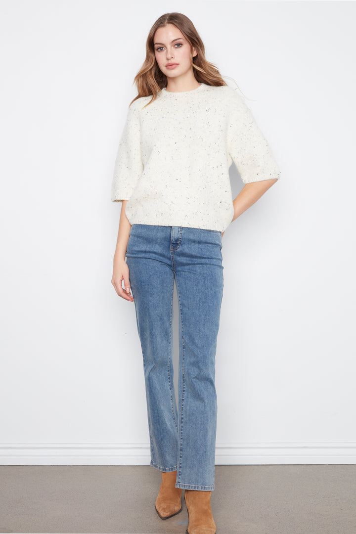 A woman modeling a cream-colored textured knit sweater with elbow-length sleeves and blue straight-leg jeans, paired with tan ankle boots, against a plain white wall.