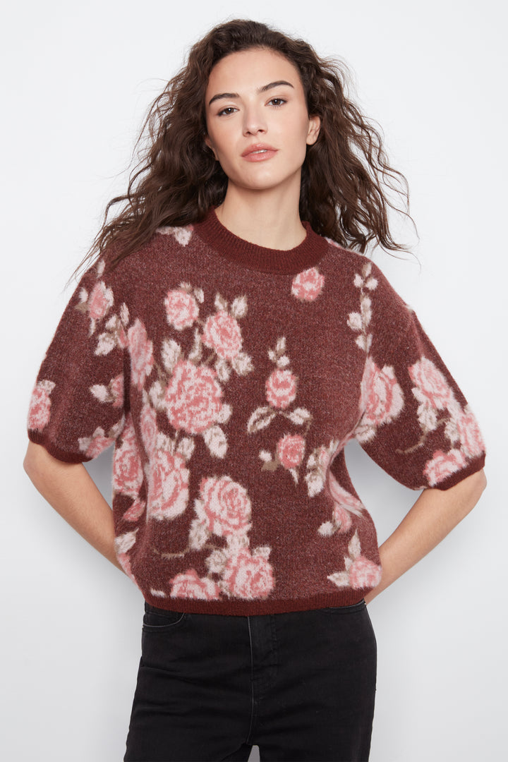 A woman wearing a short-sleeve crewneck knit sweater with a pink and white rose pattern on a dark brown background, paired with black pants, against a light gray studio background.