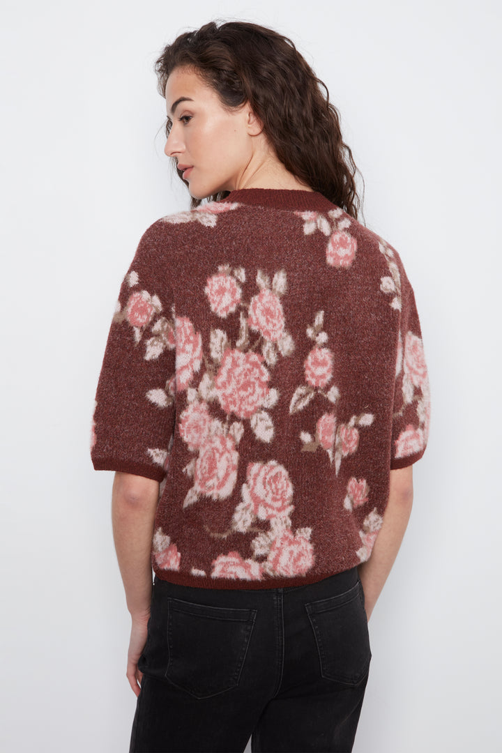 A woman shown from the back wearing a short-sleeve knit cardigan with a dusty rose floral pattern on a brown background, paired with black jeans, against a plain white studio background.