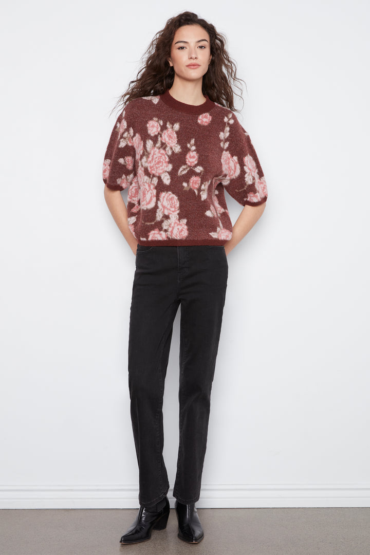 A woman modeling a short-sleeve crewneck knit sweater with a dusty rose and pink floral pattern over a dark brown base, paired with black trousers and black ankle boots, standing against a white wall.