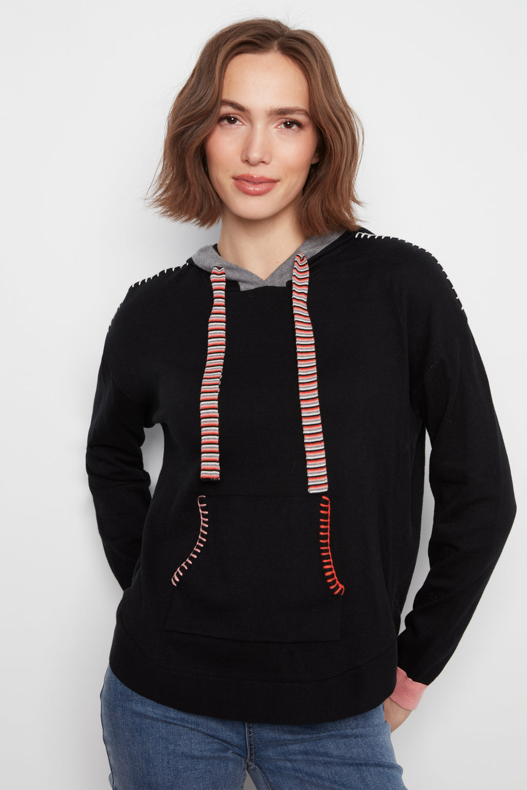 A woman wearing a black pullover hoodie with gray lining and pink-and-white striped drawstrings, paired with blue jeans, against a plain light gray background.