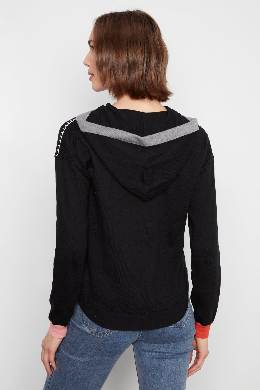A woman shown from the back wearing a black hoodie with a gray chevron pattern across the shoulders and silver chain detailing on the sleeve, paired with blue jeans against a plain white background.
