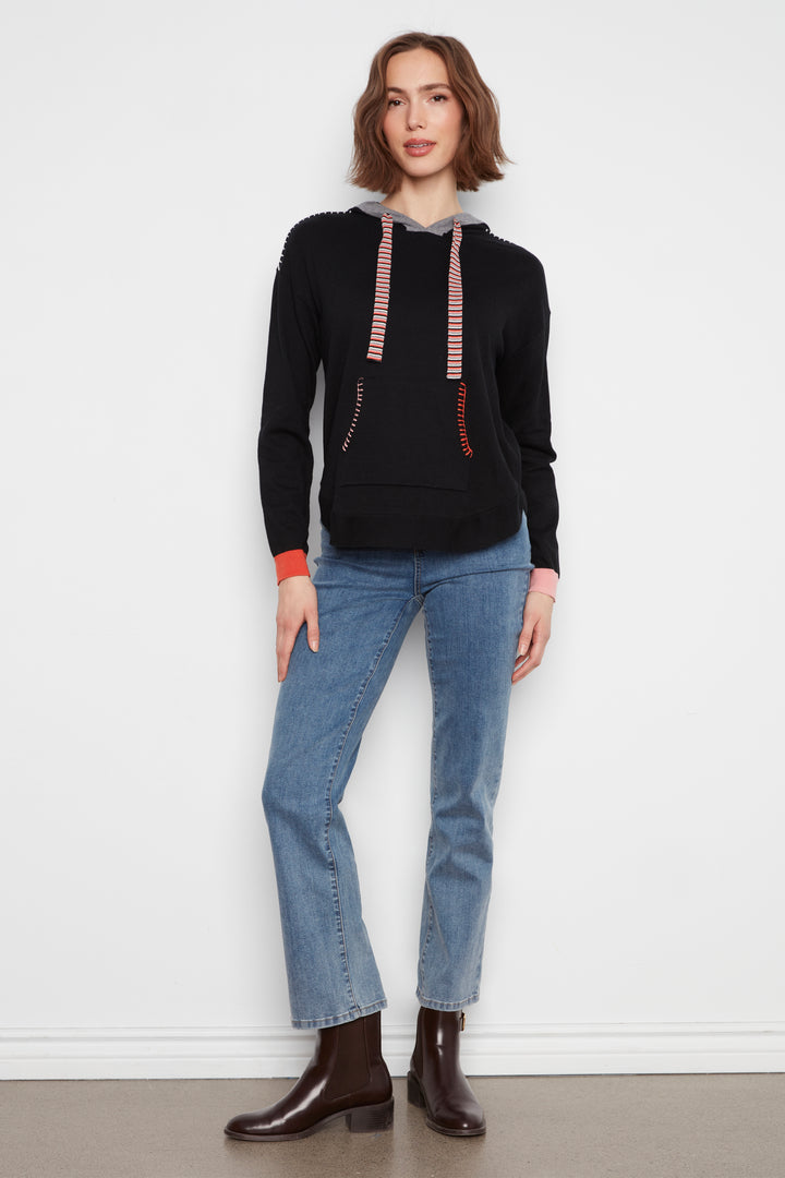 A woman modeling a black knit hoodie with pink and white striped drawstrings, paired with light blue jeans and dark brown leather ankle boots, standing against a plain white wall.