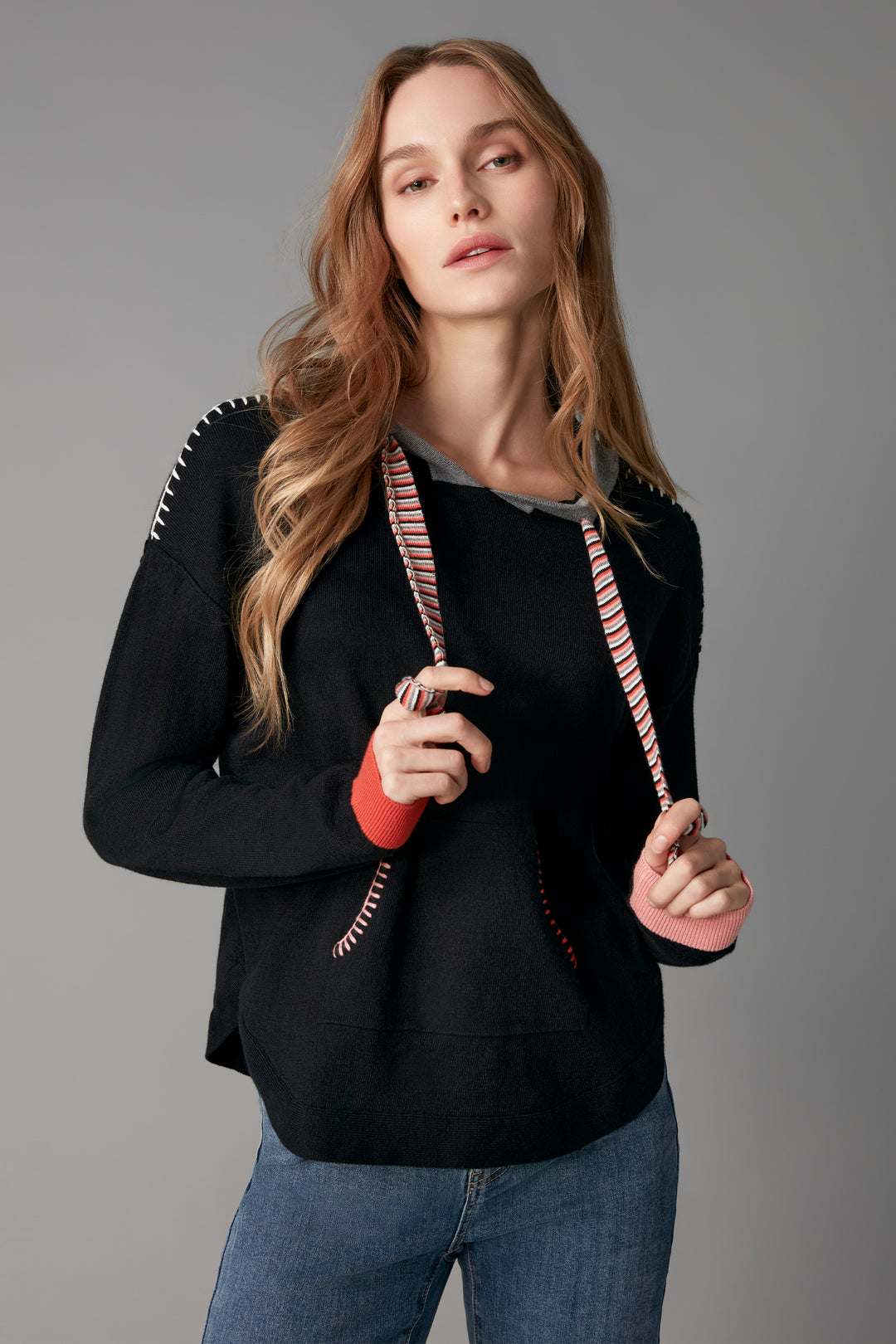 A woman modeling a black knit sweater with pink and white striped drawstrings and a gray inner collar, paired with blue jeans against a solid gray studio background.