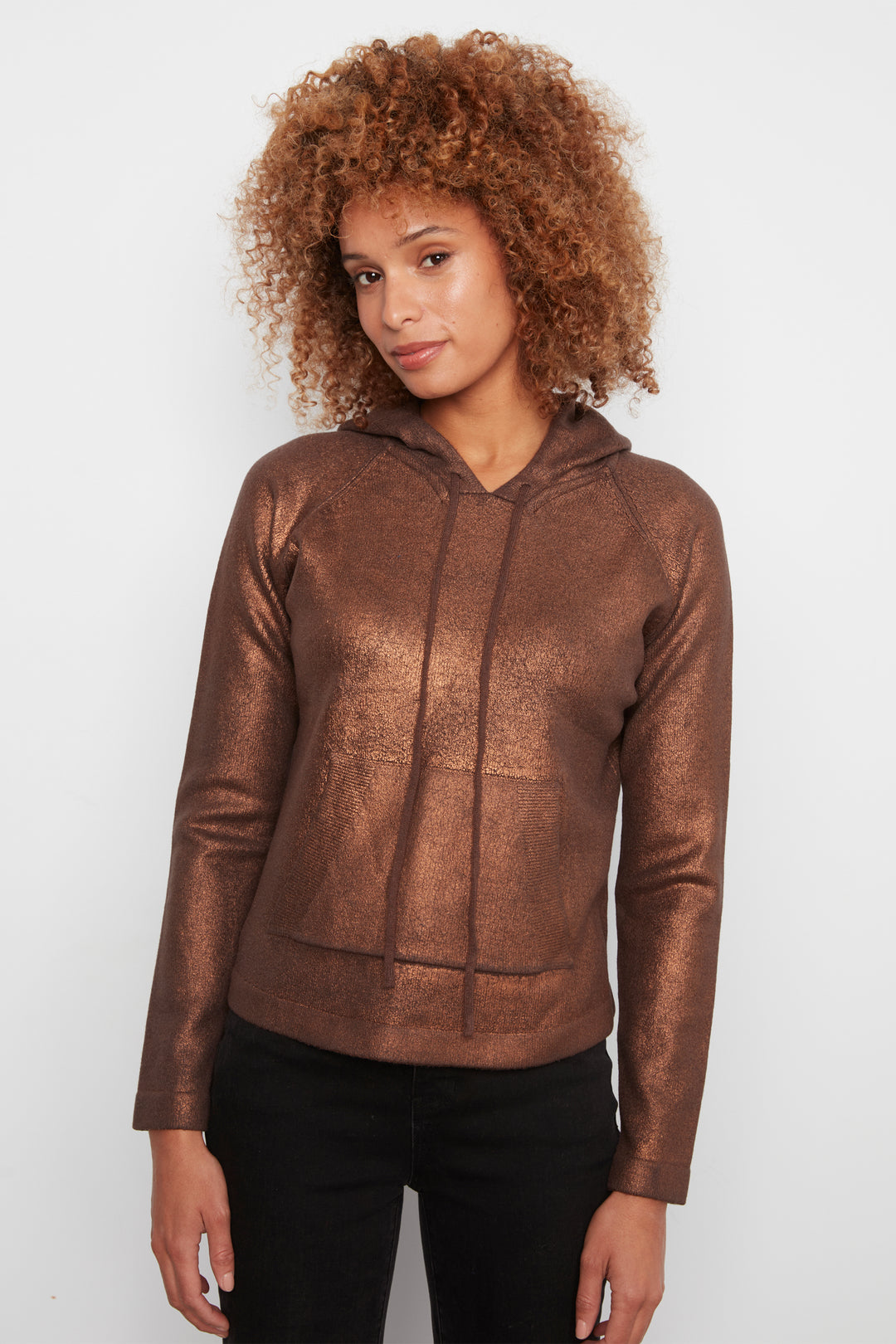 A woman with curly brown hair wearing a metallic bronze-brown hooded sweatshirt and black pants, standing against a light gray studio background.