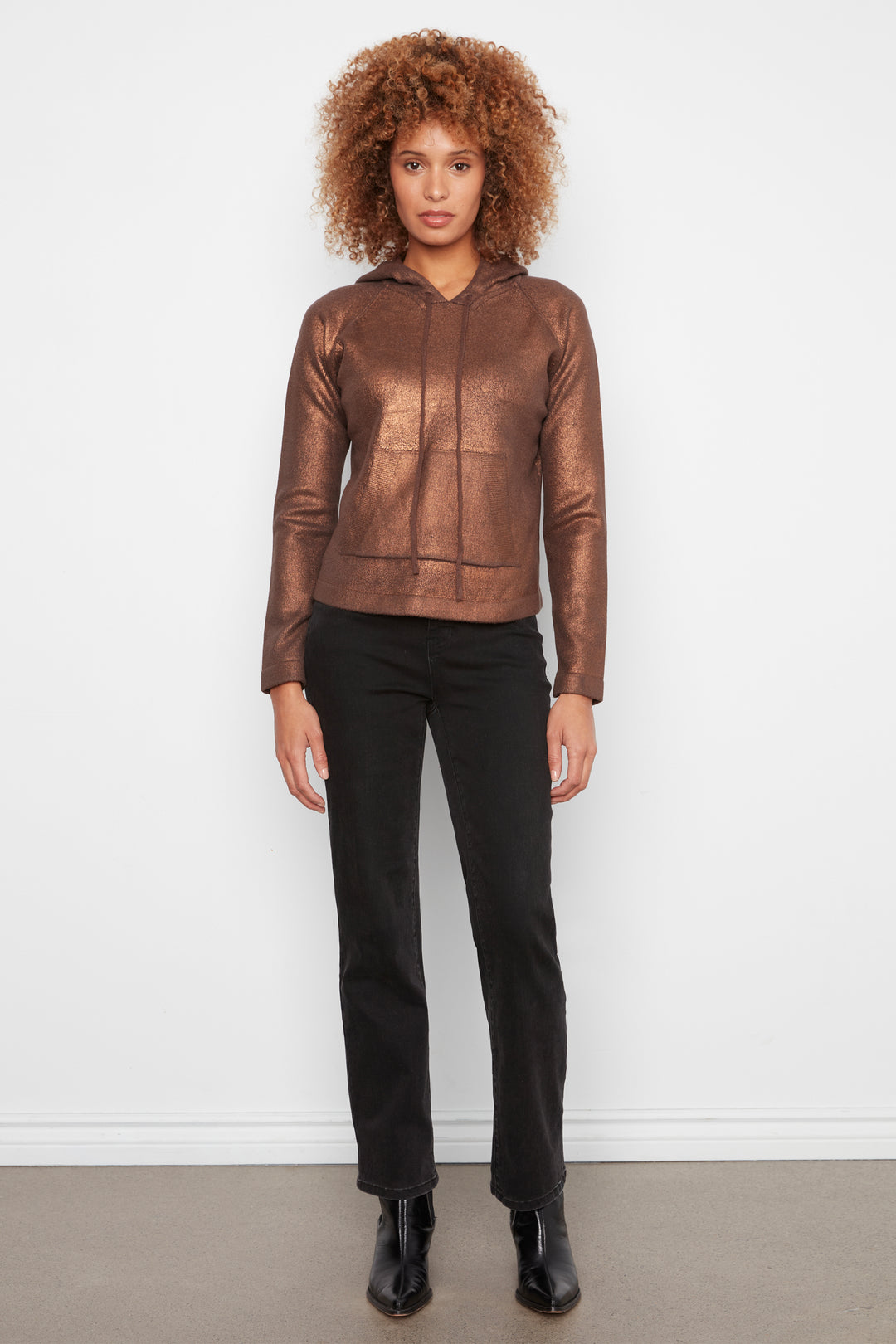 A model with curly hair wearing a metallic bronze-brown cropped hooded jacket, black pants, and black boots, standing against a plain white wall.