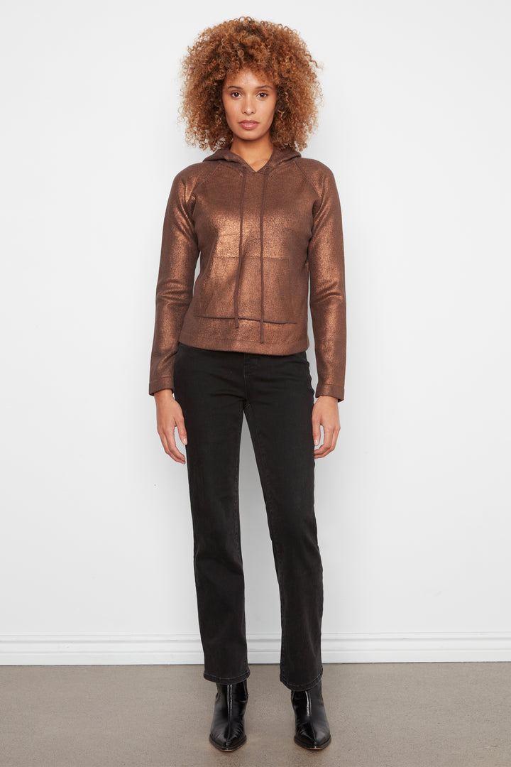 A model with curly hair wearing a metallic bronze-brown cropped hooded jacket, black pants, and black boots, standing against a plain white wall.