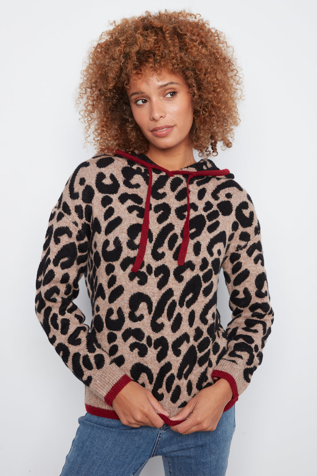 A woman with curly brown hair wearing a beige and black leopard print knit hoodie with dark red drawstrings and cuffs, paired with blue jeans, against a plain white background.