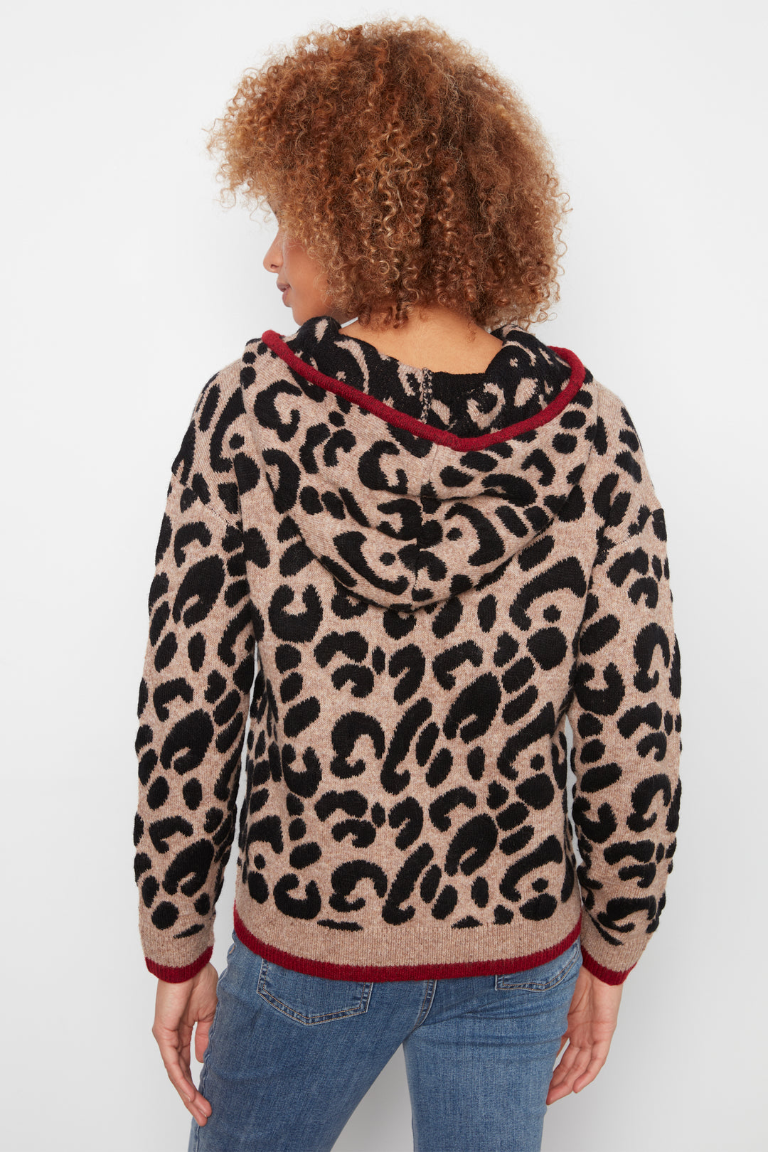 A person shown from the back wearing a beige hooded sweater with a black leopard print pattern and dark red trim, paired with blue jeans, against a plain light background.