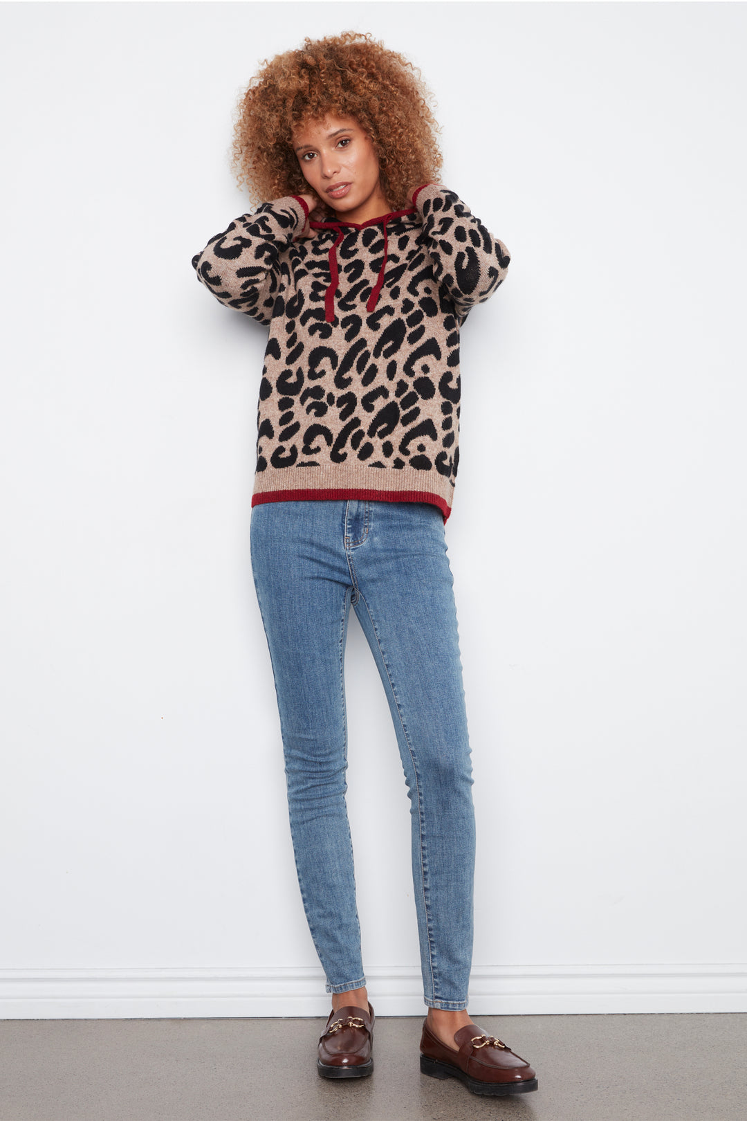 A woman with curly brown hair stands against a white wall wearing a beige and black leopard-print hoodie with red trim, blue skinny jeans, and brown loafers.